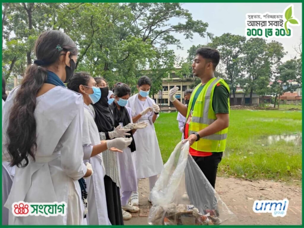 School students in uniform, wearing gloves, gathering litter into a bag alongside a volunteer on their school grounds during the 100 on 100 cleanliness drive