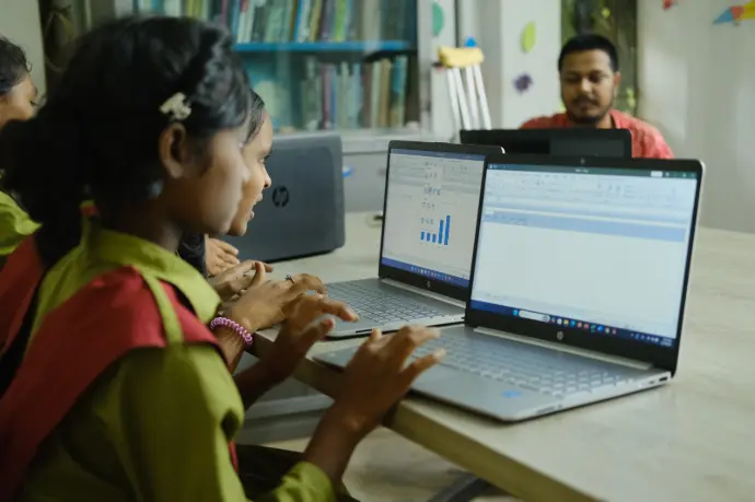 Two girls working on laptops at a digital interactive centre while a facilitator works at a desk behind them