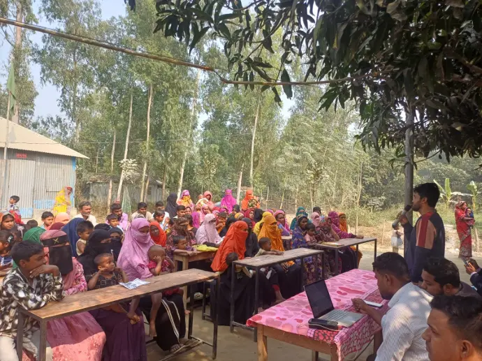 Women and children seated at benches under trees during an outdoor community session on lead exposure