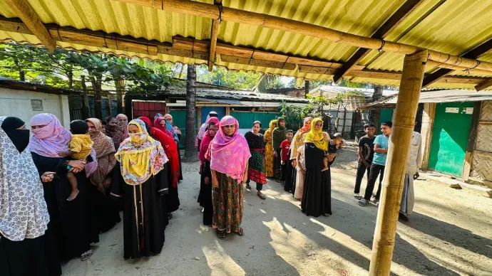 Women and children, some carrying infants, waiting in a queue under a tin shelter in a coastal neighbourhood