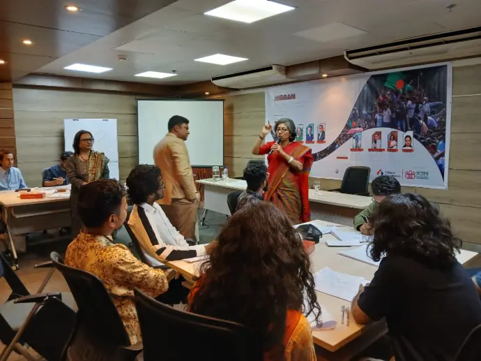 A facilitator leads a NIRBAN mental health session beside the project banner as participants take notes around a seminar table