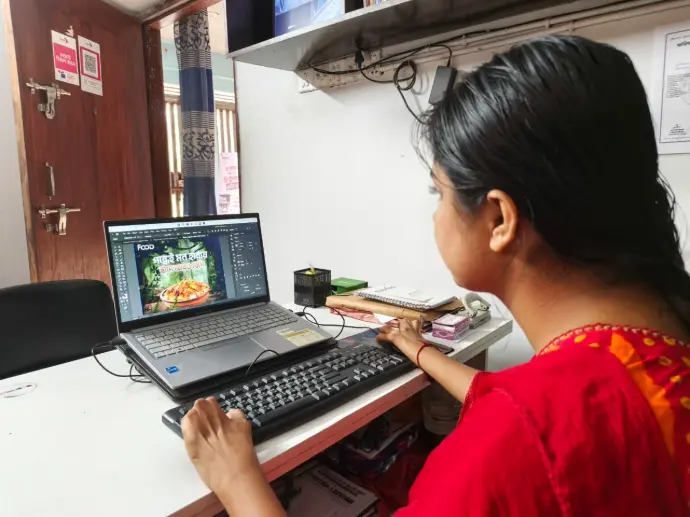 Loxme Roy seated at a computer workstation designing graphics in an office in Faridpur.